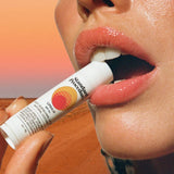 Lip protection SPF 50 balm held near glossy lips with a desert background and warm sunset lighting.