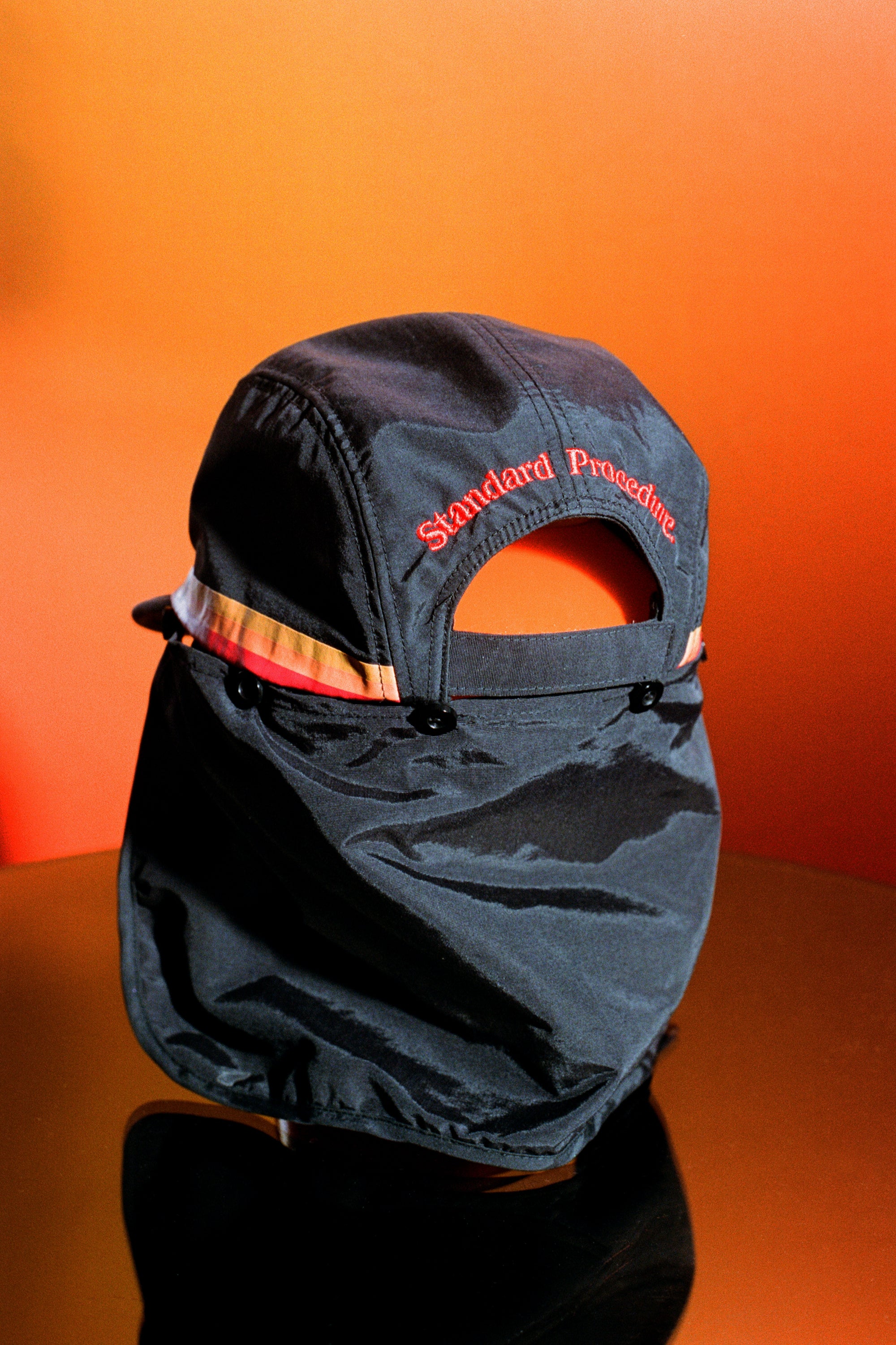 Black surf hat displayed on an orange reflective surface with detachable neck flap and embroidered Standard Procedure logo, designed for durable sun protection and outdoor wear