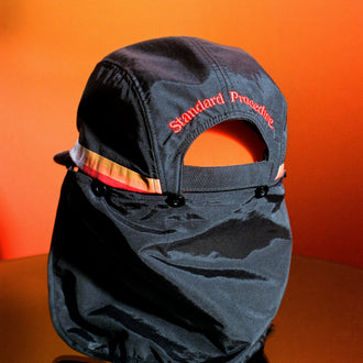 Black surf hat displayed on an orange reflective surface with detachable neck flap and embroidered Standard Procedure logo, designed for durable sun protection and outdoor wear