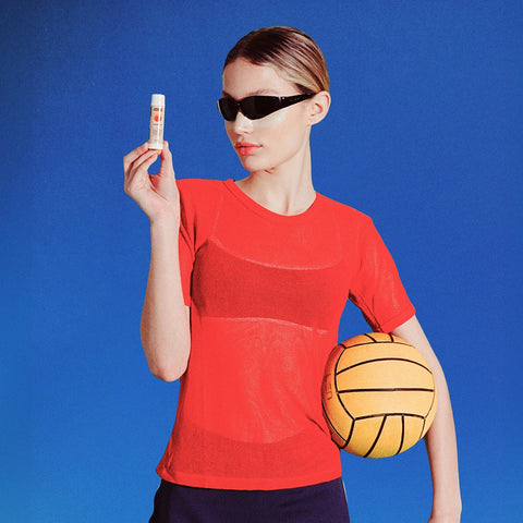 Zinc sunblock stick held up by a woman in sunglasses wearing a red top, holding a yellow volleyball against a blue background