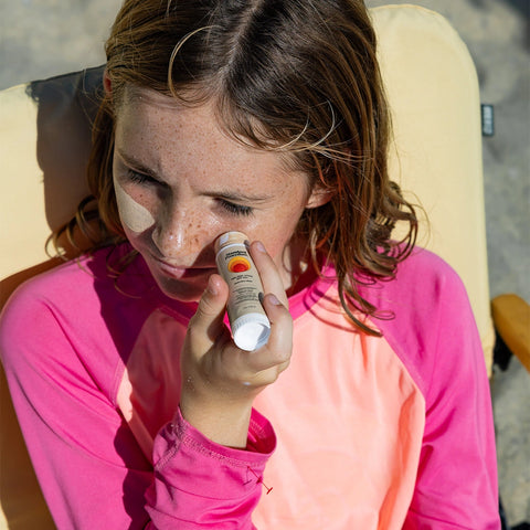Zinc sunscreen stick applied by a young girl wearing a pink rash guard while sitting outdoors, protecting her face from sun exposure