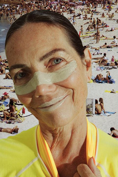 Zinc sunblock stick applied across a smiling woman’s nose & lips at a busy beach, wearing a yellow rash guard