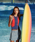 Tinted SPF 50 lip balm held by a surfer in a red-blue vest beside a rainbow surfboard with ocean waves in the background