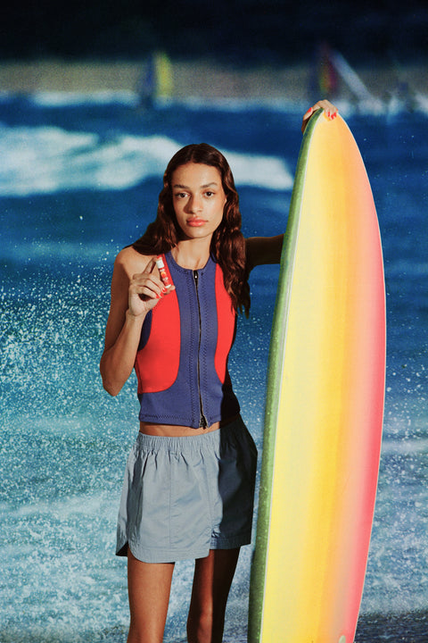 Tinted SPF 50 lip balm held by a surfer in a red-blue vest beside a rainbow surfboard with ocean waves in the background