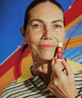 SPF 50 tinted lip balm held beside a smiling woman’s cheek, wearing a striped shirt with a colorful beach umbrella in the background