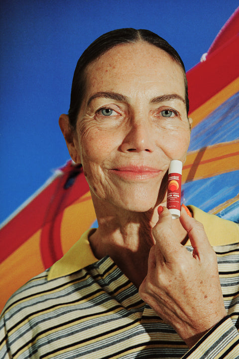 SPF 50 tinted lip balm held beside a smiling woman’s cheek, wearing a striped shirt with a colorful beach umbrella in the background