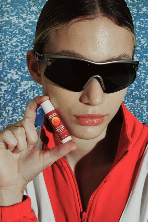 SPF 50 lip sunscreen held beside glossy lips by a person in wraparound sunglasses & a red jacket against a water-speckled background
