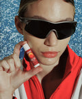 SPF 50 sunscreen for lips held beside a person’s mouth wearing wraparound sunglasses & a red jacket against a water-textured background
