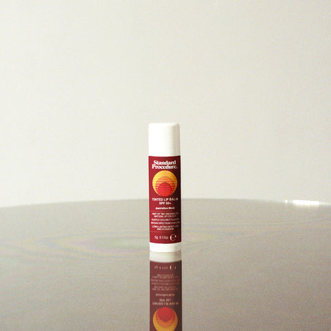 SPF 50 tinted lip balm from Standard Procedure displayed upright on a reflective surface with minimalist lighting, showcasing its Australian-made label and sun protection benefits