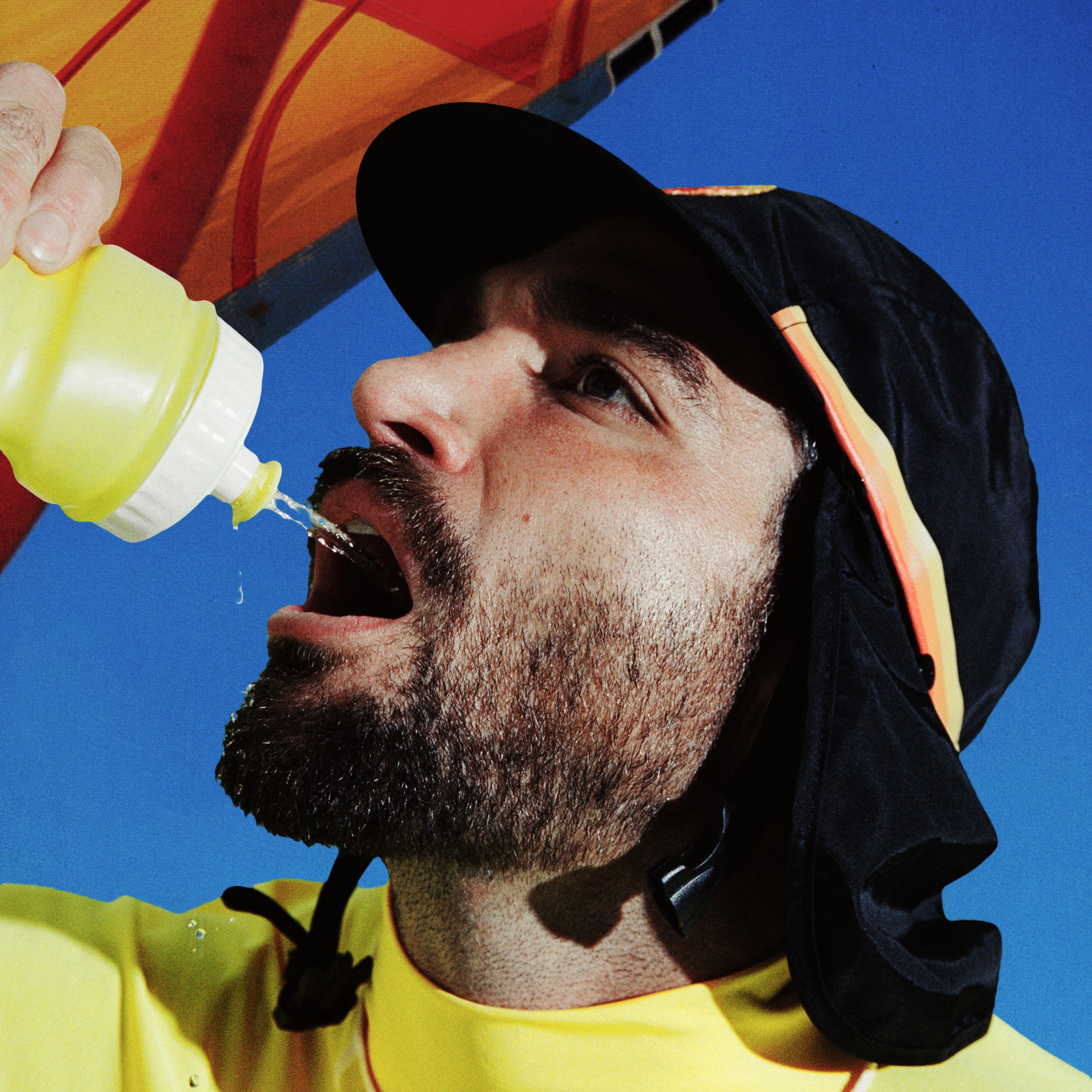 Person in black UPF 50+ flap cap sips from a water bottle under a beach umbrella.