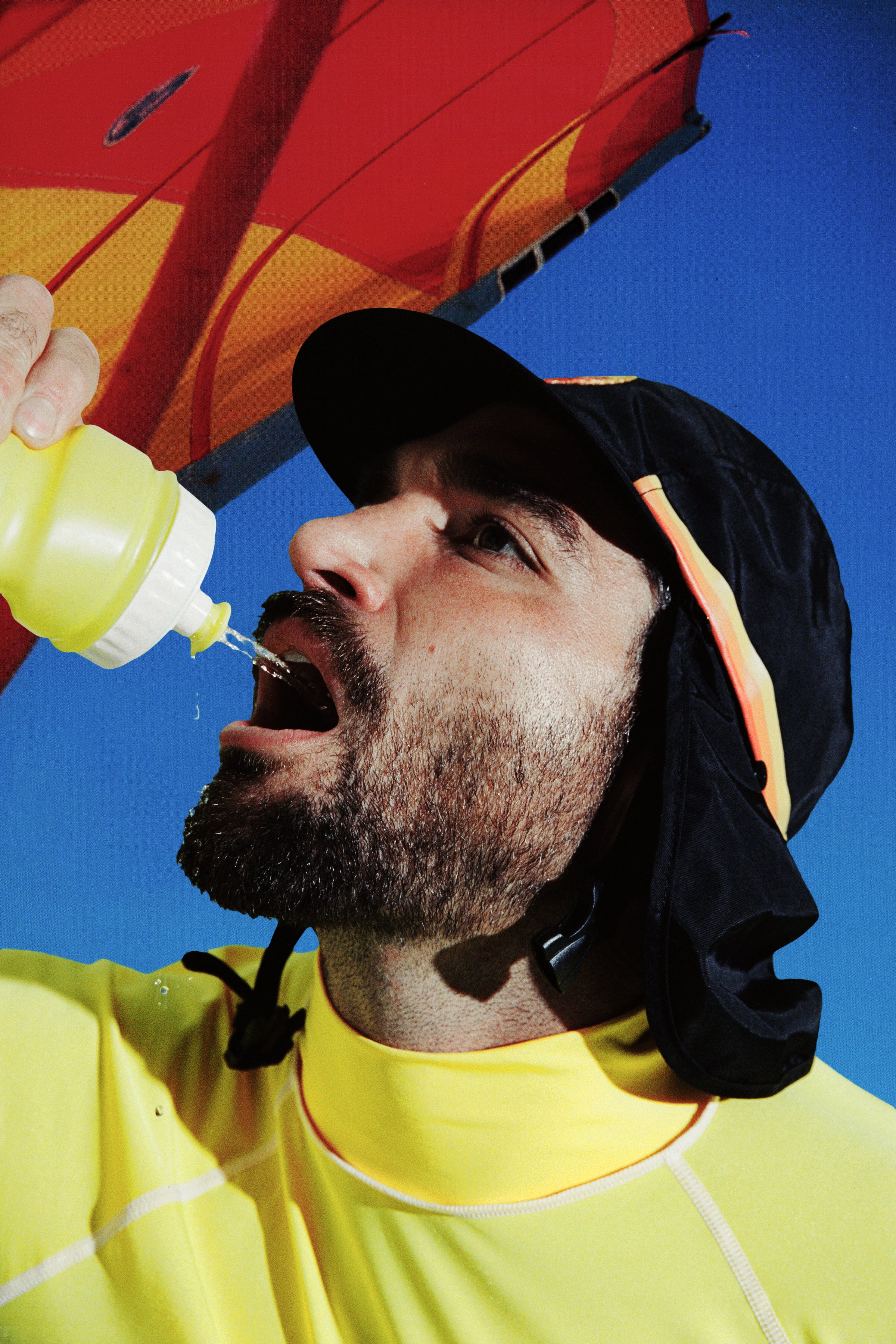 Person in black UPF 50+ flap cap sips from a water bottle under a beach umbrella.