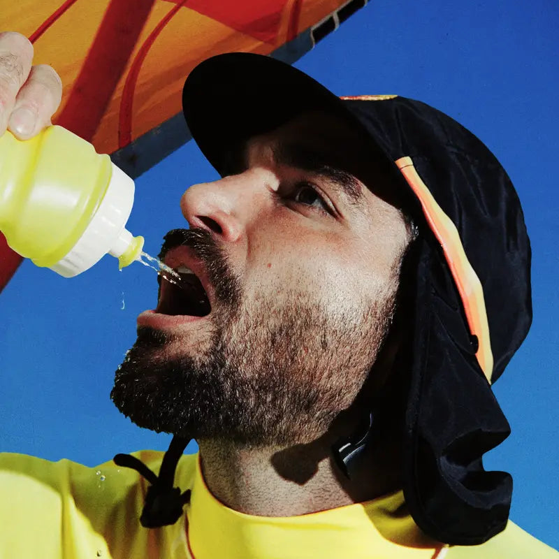 Person in black UPF 50+ flap cap sips from a water bottle under a beach umbrella.