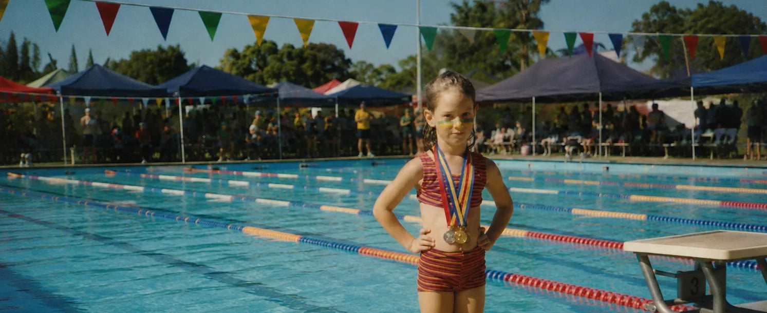 Founder image of Zepha Jackson as Child with swimming medals standing by a public pool with flags and spectators in the background wearing Standard Procedure Colored SPF 50+ Zinc Stripes on her face