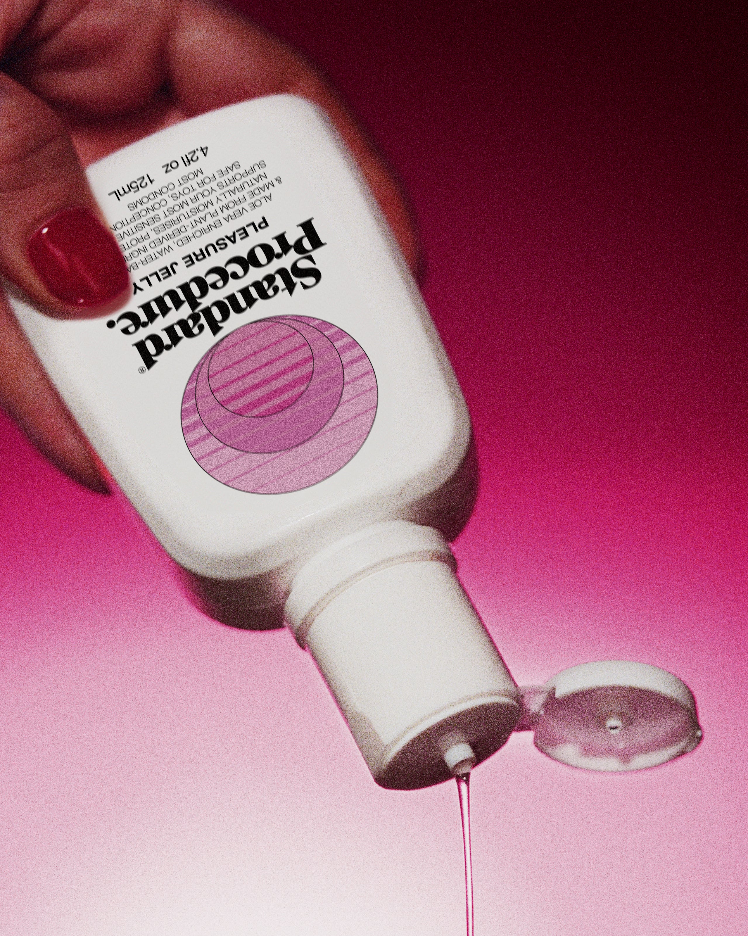 Red-manicured hand squeezing Pleasure Jelly aloe vera lubricant bottle, clear gel streaming onto pink surface.