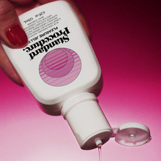 Red-manicured hand squeezing Pleasure Jelly aloe vera lubricant bottle, clear gel streaming onto pink surface.