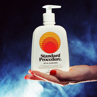 SPF 50 sunscreen pump bottle held in a hand against a smoky blue backdrop.