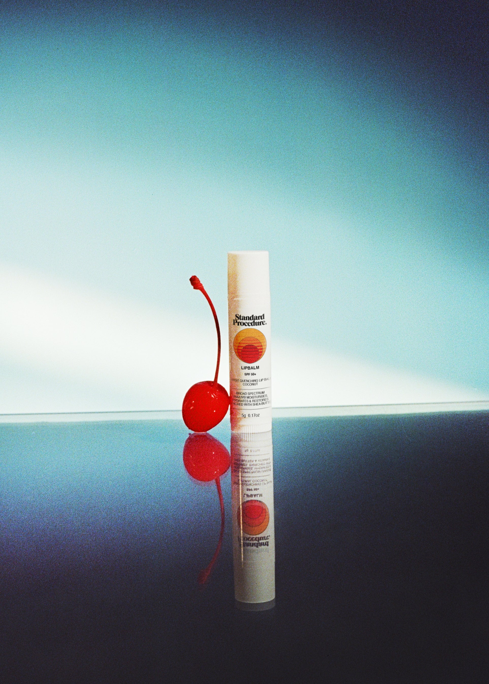 Lip protection SPF 50 balm displayed beside a red cherry on a reflective surface with a blue gradient background.