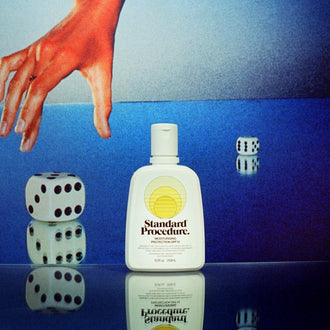 Standard Procedure SPF 15 moisturiser bottle on a mirrored surface beside rolling dice and a sunburnt hand.