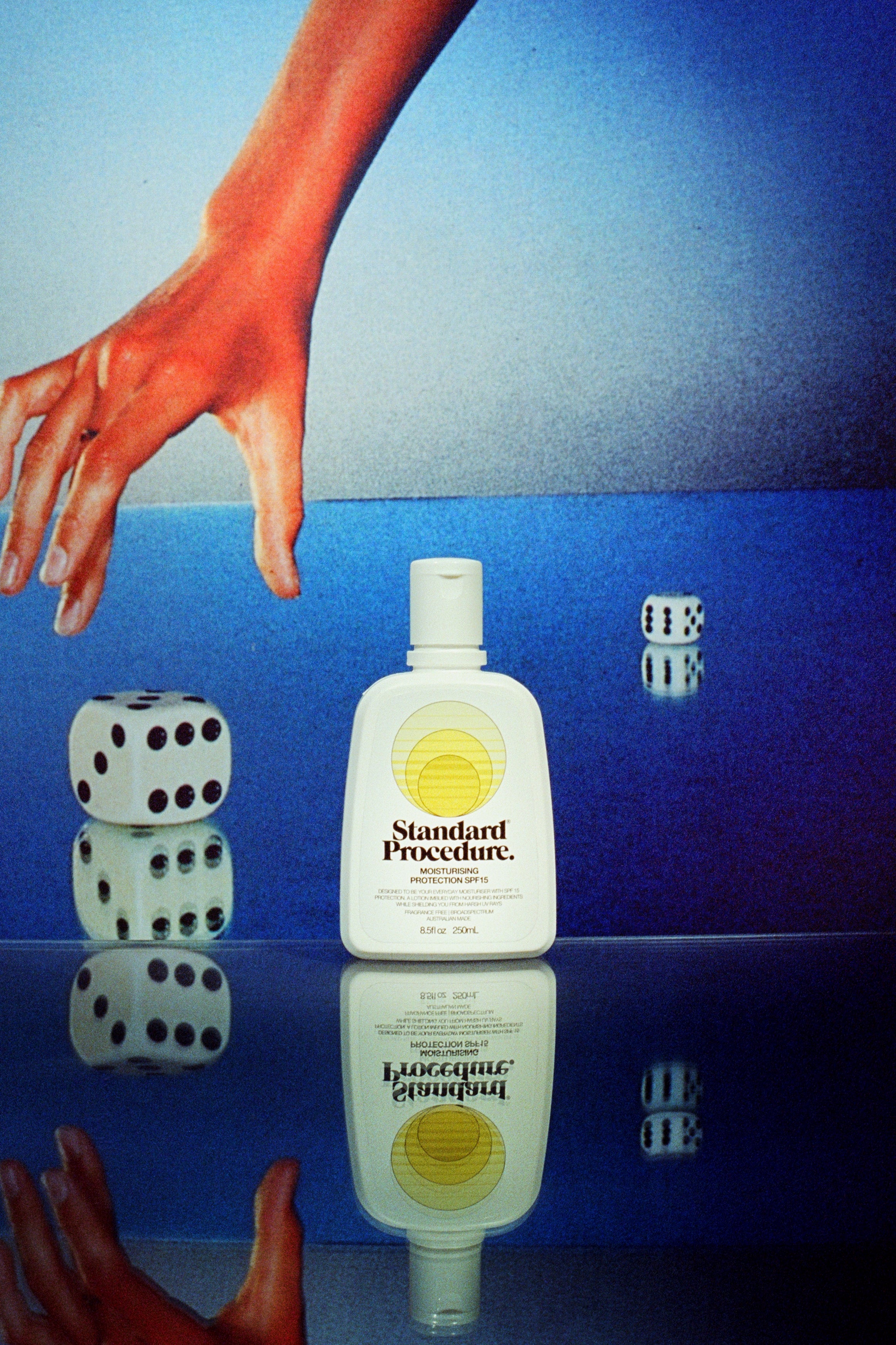 Standard Procedure SPF 15 moisturiser bottle on a mirrored surface beside rolling dice and a sunburnt hand.