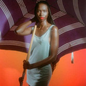 Editorial sun protection campaign for Standard Procedure SPF 15 moisturiser, model shaded under a striped parasol.