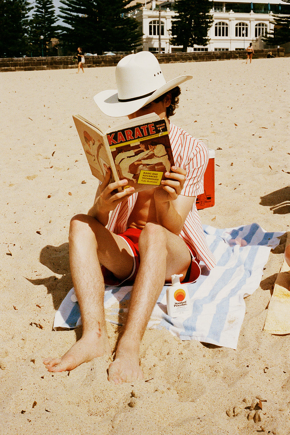 Beachgoer in cowboy hat reads a karate book beside a Standard Procedure SPF 50+ sunscreen bottle on a striped towel.