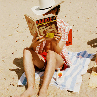 Beachgoer in cowboy hat reads a karate book beside a Standard Procedure SPF 50+ sunscreen bottle on a striped towel.