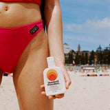 mineral sunscreen broad spectrum spf 50 bottle held by a beachgoer in a red swimsuit against a sunlit sandy beach backdrop.