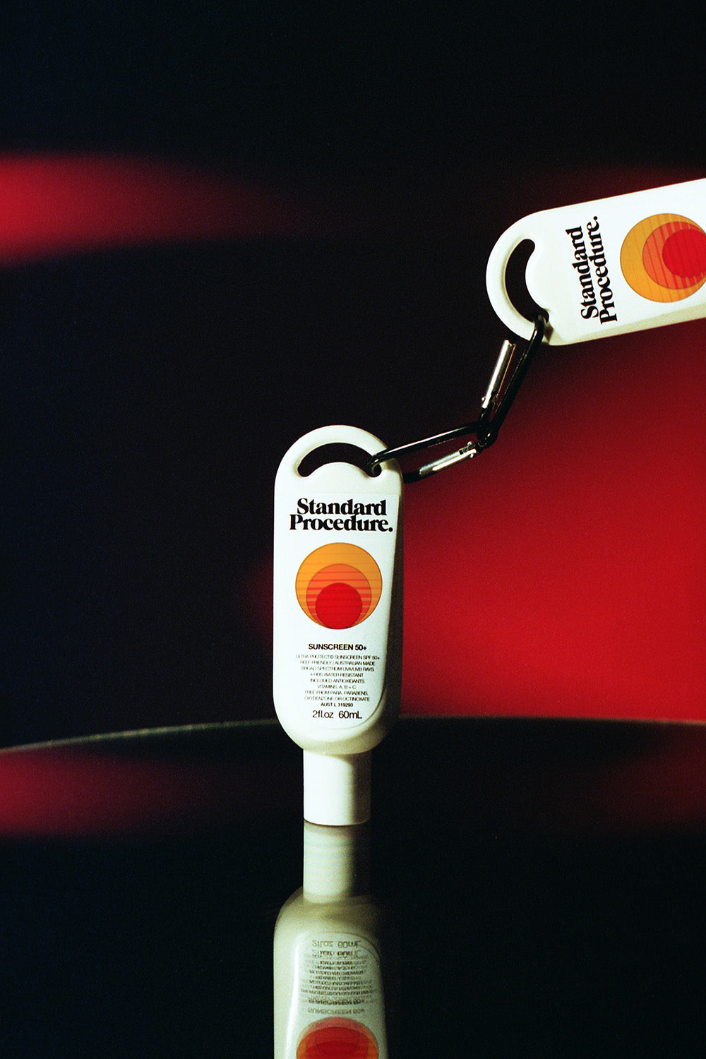 Travel size SPF 50 sunscreen from Standard Procedure connected by a black carabiner, displayed against a red background to highlight its portable design and strong sun protection formula