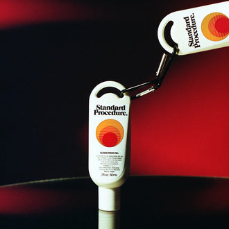 Travel size SPF 50 sunscreen from Standard Procedure connected by a black carabiner, displayed against a red background to highlight its portable design and strong sun protection formula