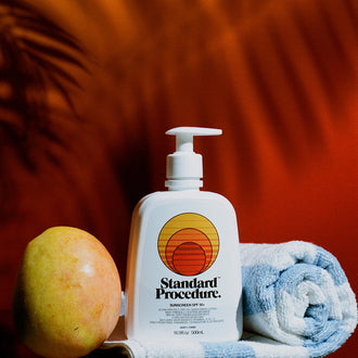 Sunblock SPF 50 pump bottle beside a fresh mango and rolled beach towel on a reflective surface.