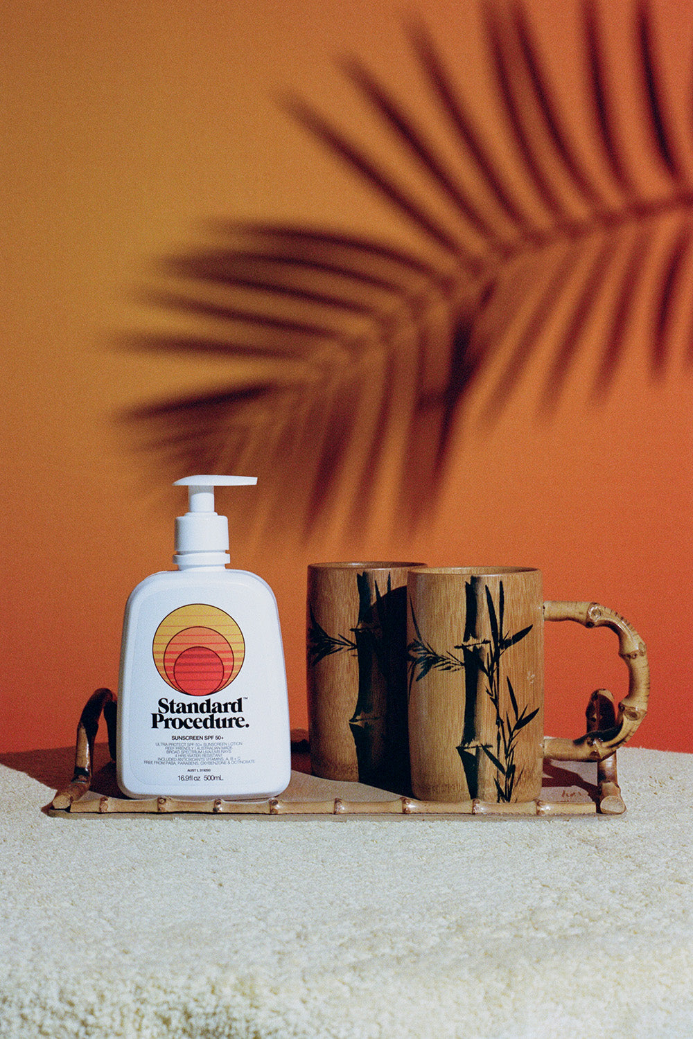 Sun block 50 SPF bottle by Standard Procedure placed on a bamboo tray with wooden mugs against a tropical sunset background.