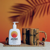 Sun block 50 SPF bottle by Standard Procedure placed on a bamboo tray with wooden mugs against a tropical sunset background.