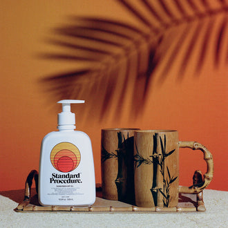 Sun block 50 SPF bottle by Standard Procedure placed on a bamboo tray with wooden mugs against a tropical sunset background.