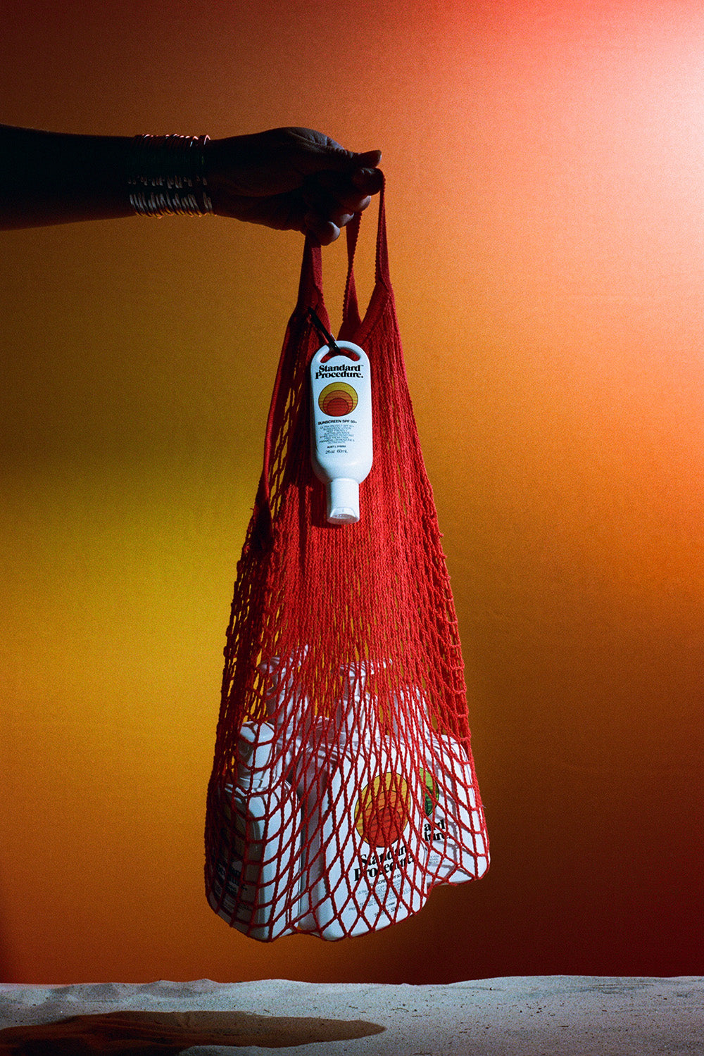 Australian sunscreen from Standard Procedure displayed in a red mesh bag held by a hand, set against a warm orange gradient background, showcasing portable SPF protection for beach days