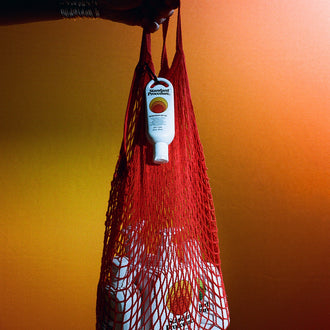 Australian sunscreen from Standard Procedure displayed in a red mesh bag held by a hand, set against a warm orange gradient background, showcasing portable SPF protection for beach days