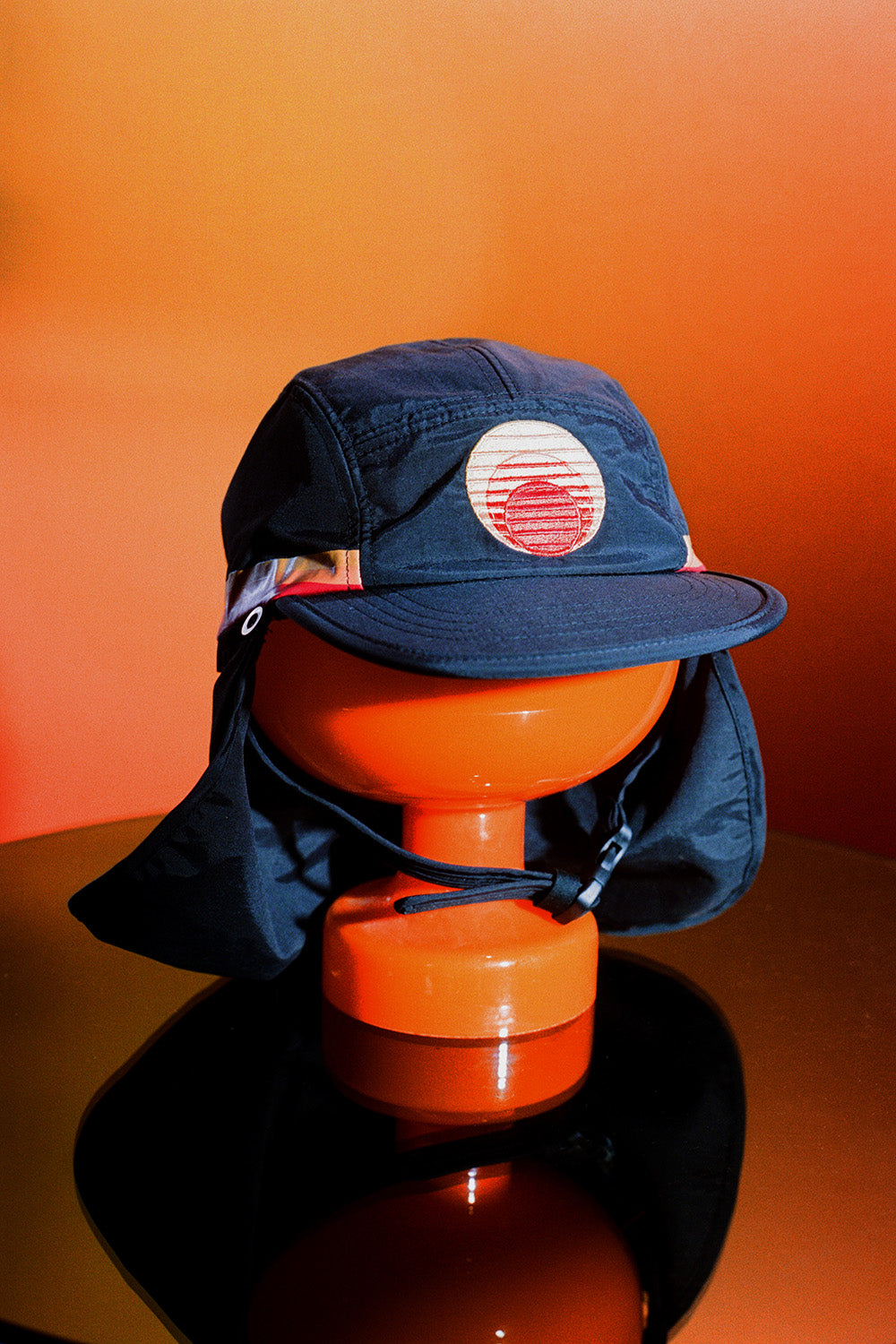 Navy UPF 50+ sports hat with neck flap and sunset patch on orange display stand.