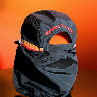 Rear view of black Standard Procedure flap cap with neck cover against an orange backdrop.