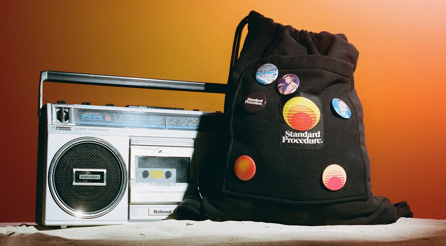 Black Standard Procedure towel bag with retro badges beside a vintage boombox on sand.