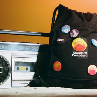 Black Standard Procedure towel bag with retro badges beside a vintage boombox on sand.