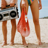 Sun cream SPF 50 clipped to a red mesh beach bag held by a woman standing barefoot on the sand.