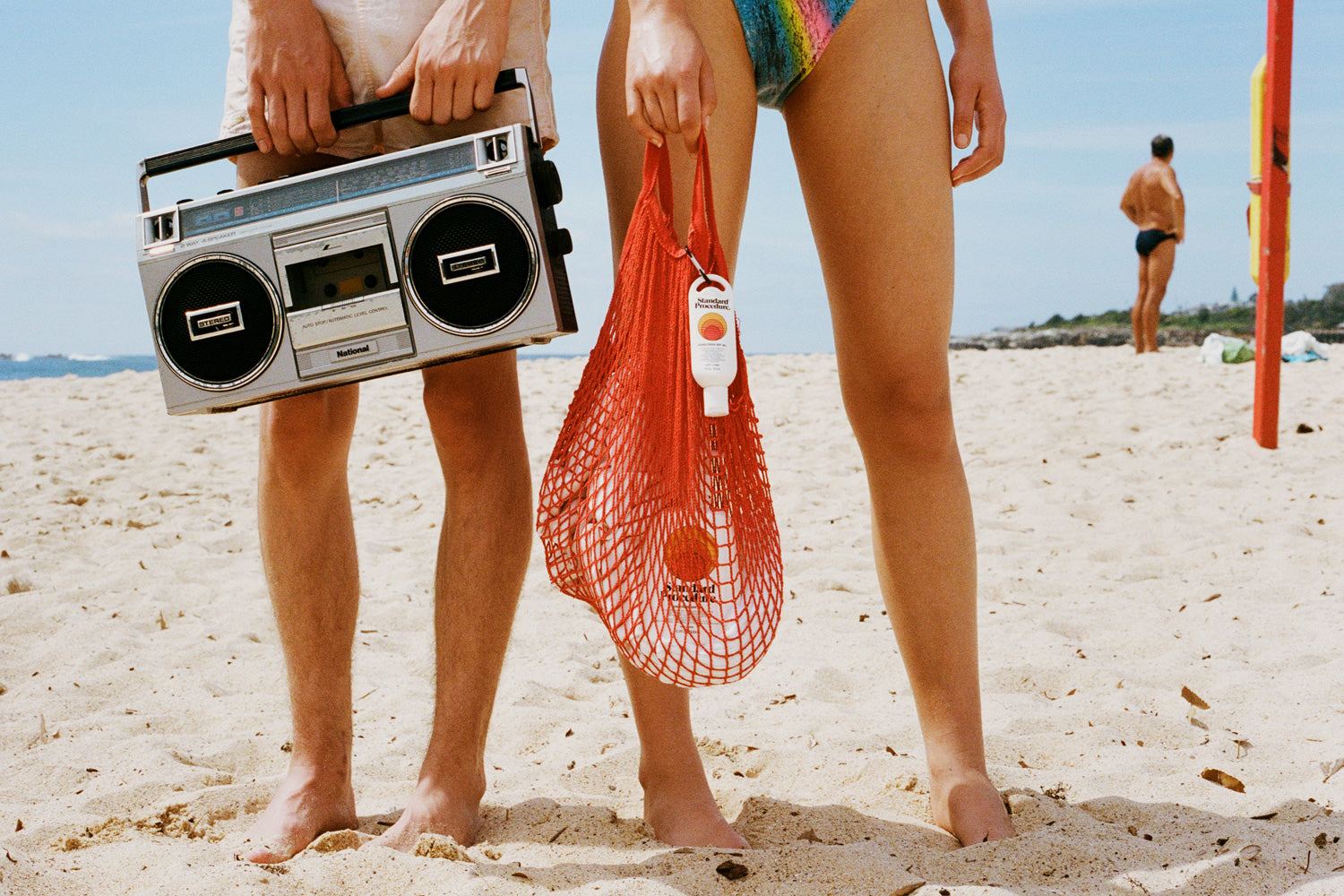 Sun cream SPF 50 clipped to a red mesh beach bag held by a woman standing barefoot on the sand.