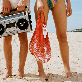 Sun cream SPF 50 clipped to a red mesh beach bag held by a woman standing barefoot on the sand.