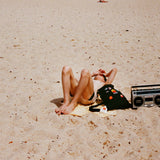 Sunbather on sandy beach with Standard Procedure SPF 50+ sunscreen bottle, backpack, and vintage boombox.
