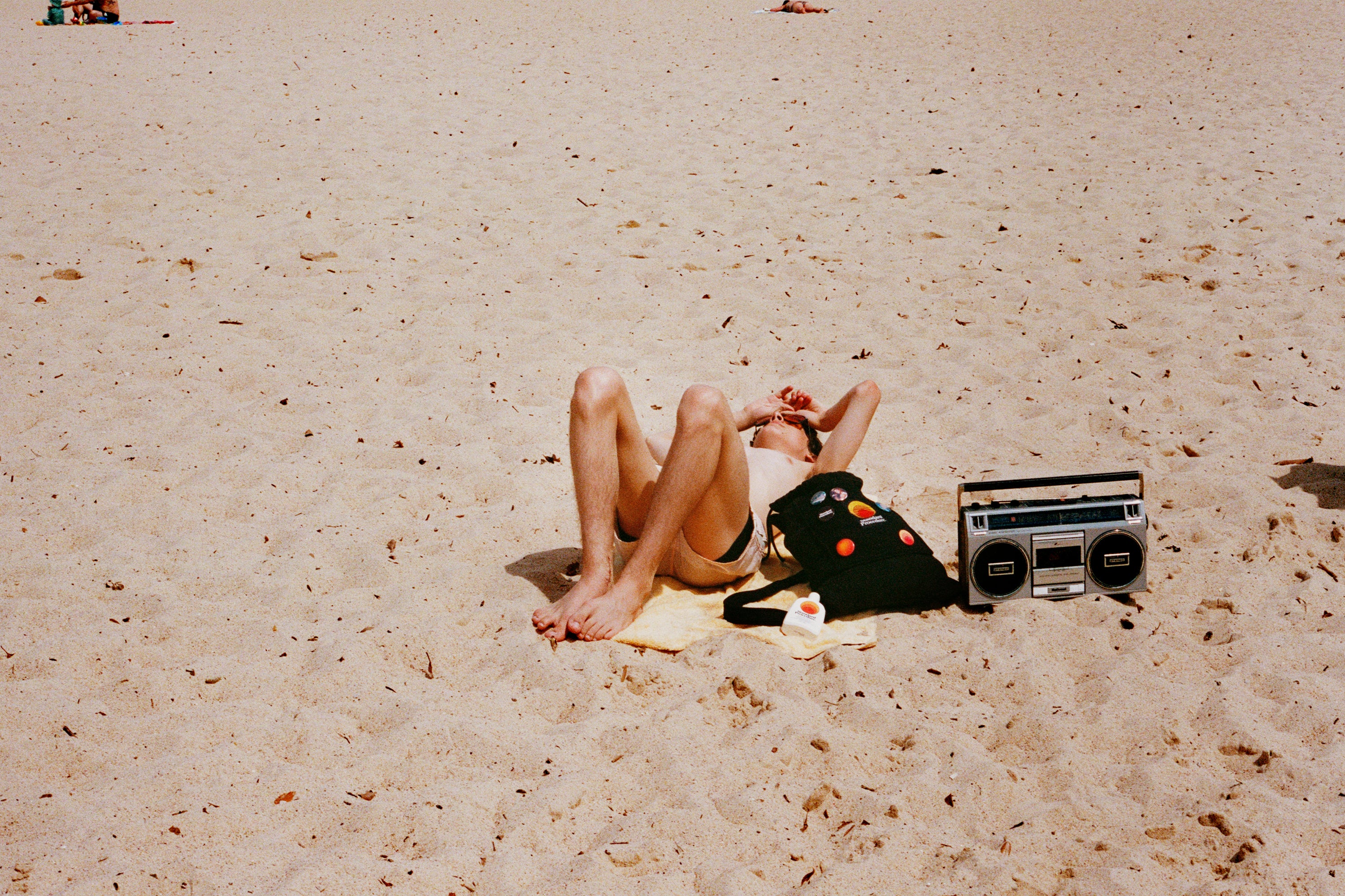Sunbather on sandy beach with Standard Procedure SPF 50+ sunscreen bottle, backpack, and vintage boombox.