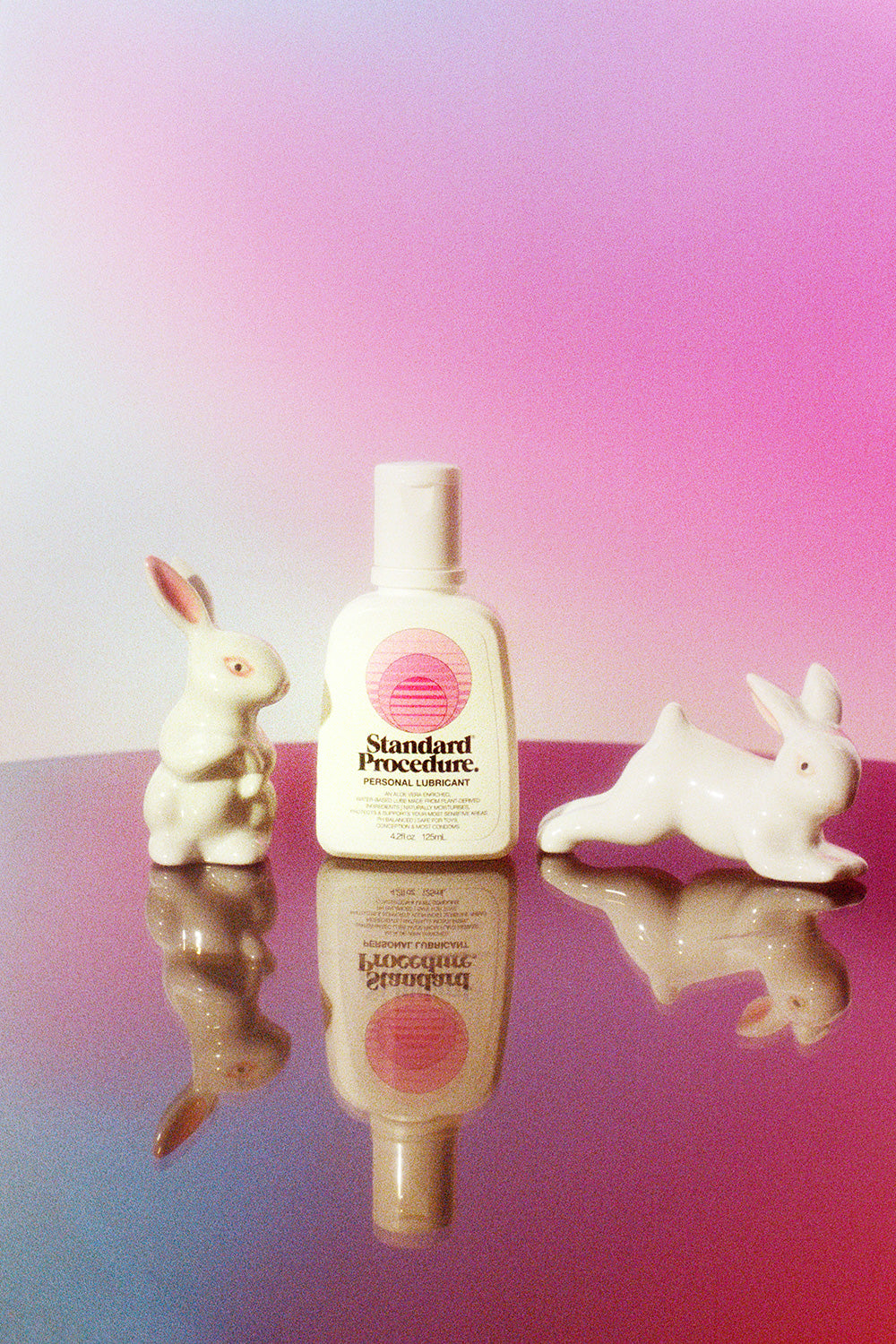 Aloe vera Pleasure Jelly lubricant bottle flanked by two ceramic rabbits on a mirrored pink surface.
