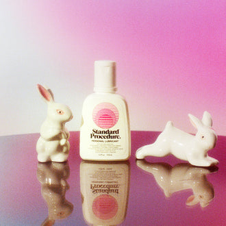 Aloe vera Pleasure Jelly lubricant bottle flanked by two ceramic rabbits on a mirrored pink surface.