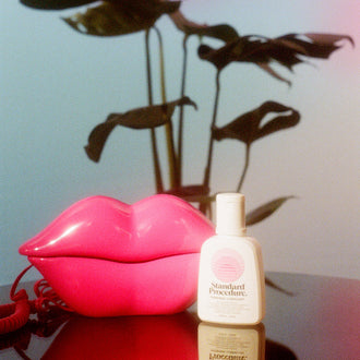 Standard Procedure Pleasure Jelly lubricant bottle beside a pink lips telephone and monstera plant.