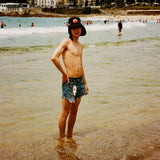 Black UPF 50+ sports hat with neck flap worn at a sunny beach shoreline.
