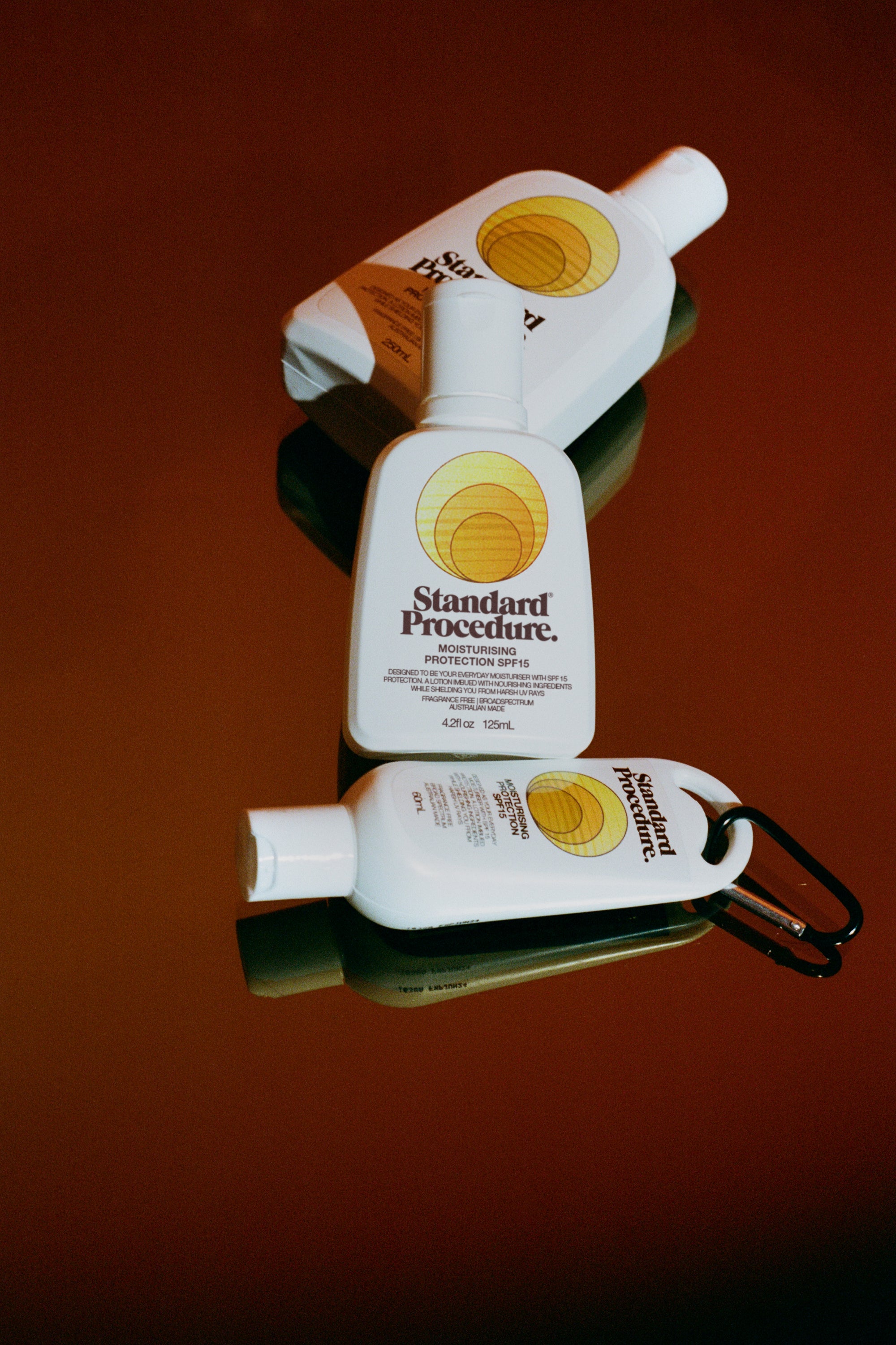 Three Standard Procedure SPF 15 moisturising sunscreen bottles in varying sizes on deep red surface.