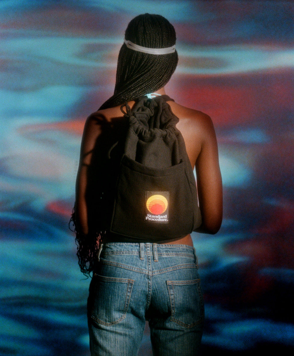 Black Standard Procedure towel bag worn as a drawstring backpack against a sunset-toned backdrop.